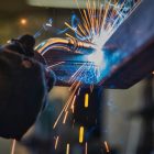 Closeup view of a gloved hand welding a joint between two pieces of metal with sparks emanating from the contact point
