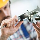 A worker with his face blurred in the background and his hands clear in the foreground using a mechanical measuring instrument on a fabricated circular metal piece